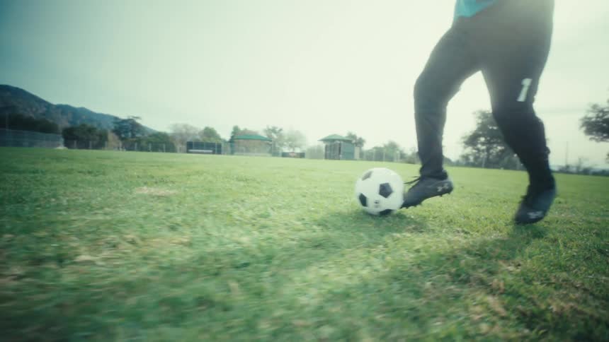 A view from ground level shows the legs and feet of a soccer player as he uses his feet to dribble a soccer ball around yellow poles on a green field