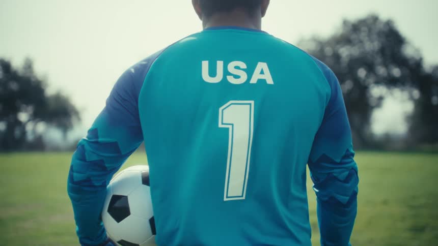 A young man wearing a blue jersey and goalie gloves squats on a green field next to a soccer ball