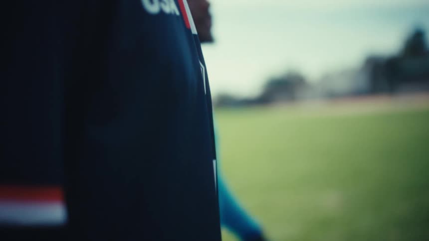 Two soccer players wearing usa jerseys walk across a grass field under a bright sky one player carries a soccer ball at his side while the camera follows them from a close low-angle perspective