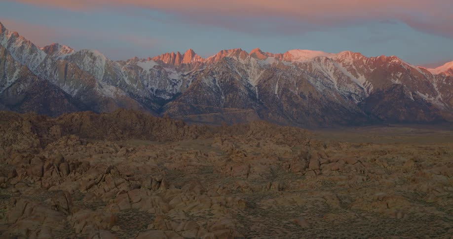 An aerial shot glides over a rocky desert landscape scattered with large bouldersin the distance a massive range of snowcapped mountains is illuminated by the warm orange glow of a setting sun