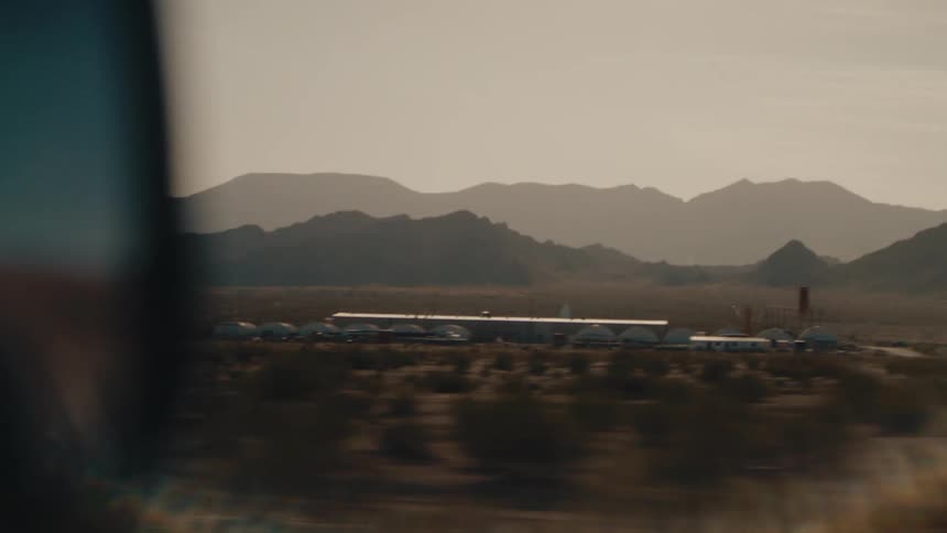 A desert landscape from the perspective of a moving vehicle with rugged mountains silhouetted against a hazy lateafternoon sky in the foreground a blurred sideview mirror and lens flare frame a passing cluster of lowprofile bui