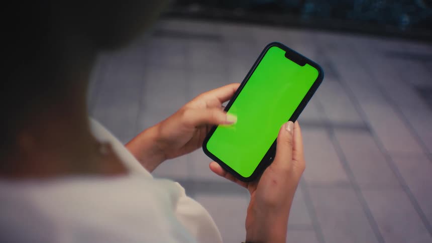 A person stands outdoors in the evening and holds a smartphone with a green screen in a close view while people walk past them on a paved walkway in the background