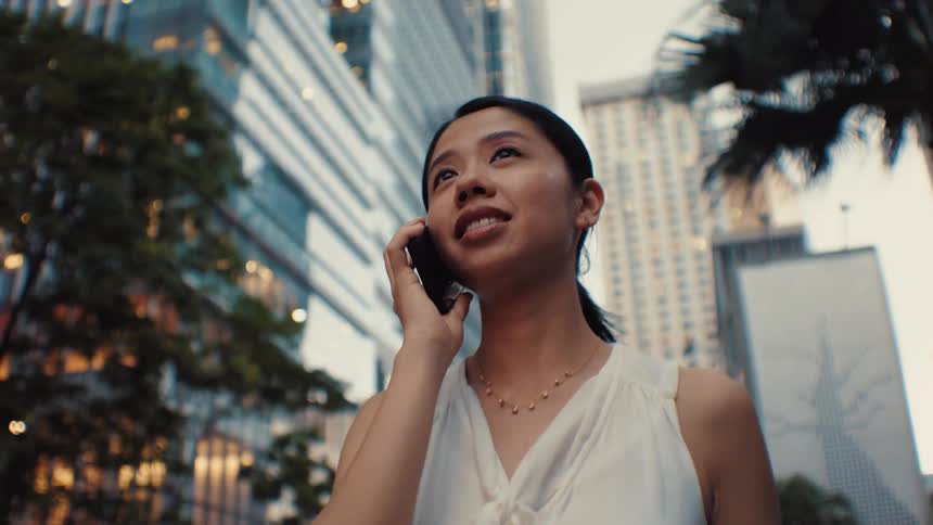 A woman wearing a white sleeveless top walks through a city with tall buildings while talking on a mobile phone and smiling in a low view