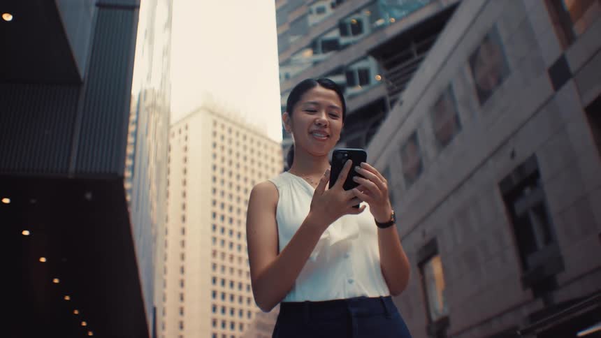 A woman wearing a white sleeveless top and dark trousers walks through a city with tall buildings stops to check her phone while smiling then puts the device in her pocket and continues walking