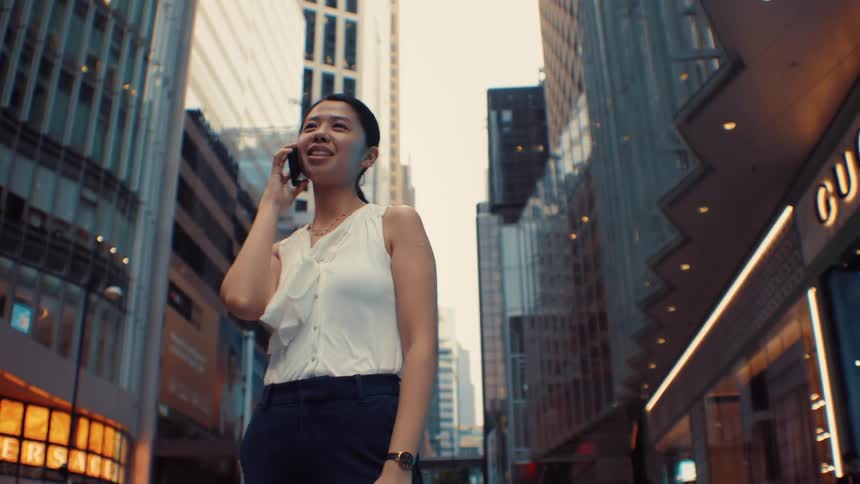 A woman in business attire stands on a city street between tall buildings smiling as she talks on her phone beside a passing doubledecker bus