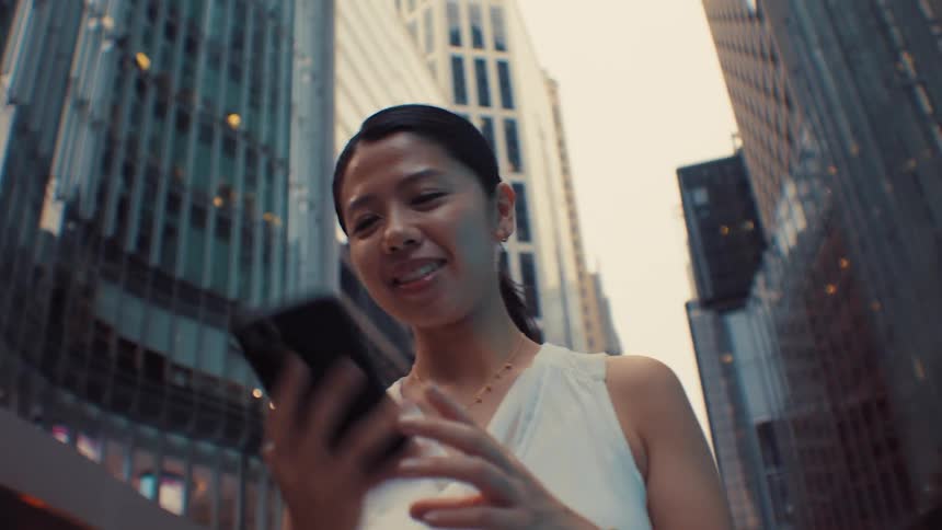 A smiling asian woman walks through a dense city district while talking on her smartphone