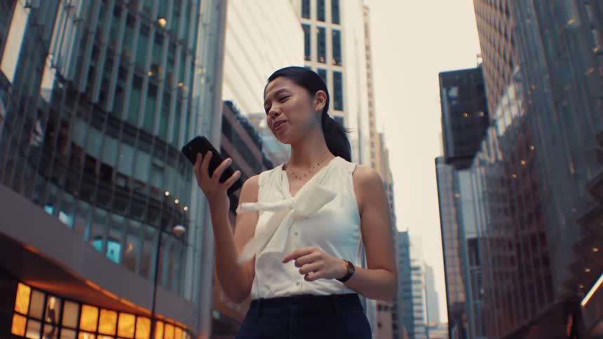 A woman wearing a white sleeveless top and dark pants walks through a city and smiles while talking on her phone with tall modern buildings and city traffic in the background