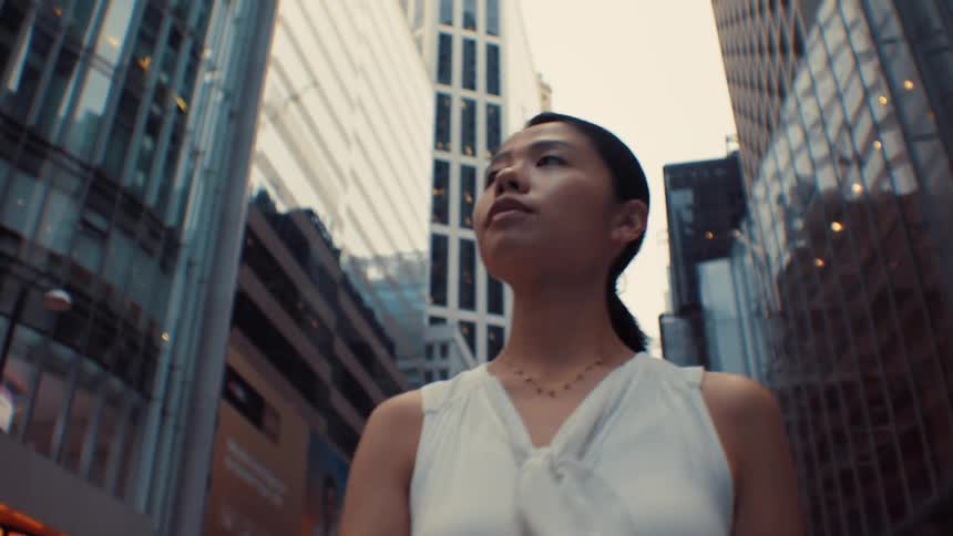 A woman in a white sleeveless blouse walks confidently through a canyon of tall glass skyscrapers in a modern city