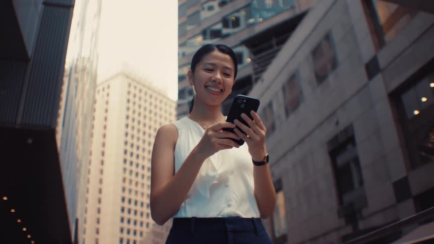 A woman wearing a white sleeveless top and dark pants walks through a city and smiles while looking at her phone with tall buildings and a moving bus in the background