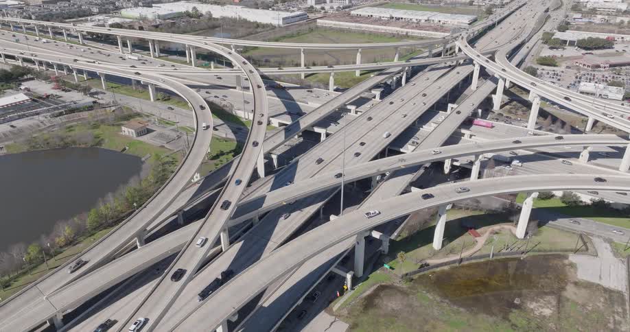 A view from above shows many cars and trucks driving along intersecting concrete highway ramps and overpasses on a bright sunny day