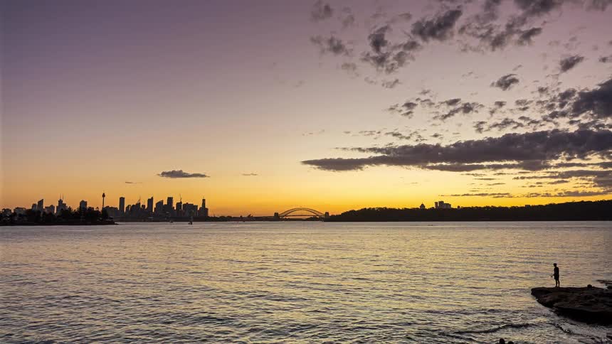 A wide view shows the sun setting over a city skyline across a body of water as boats pass by and the sky changes from orange to dark blue while city lights begin to glow and reflect on the surface