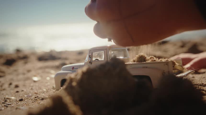 A closeup shot shows a toy pickup truck on the beach as a hand presses sand into its bed with the ocean blurred in the background