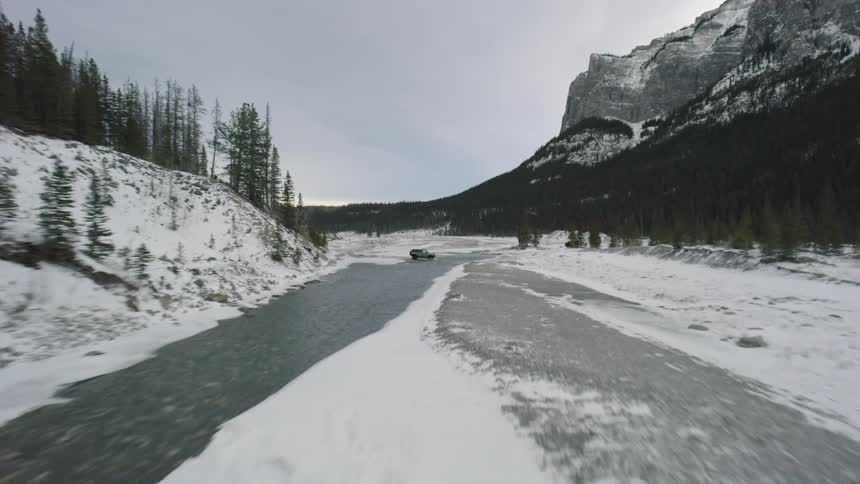 A dark truck drives across a snowy and rocky riverbank next to a flowing stream in a valley surrounded by evergreen trees and large mountains under an overcast sky