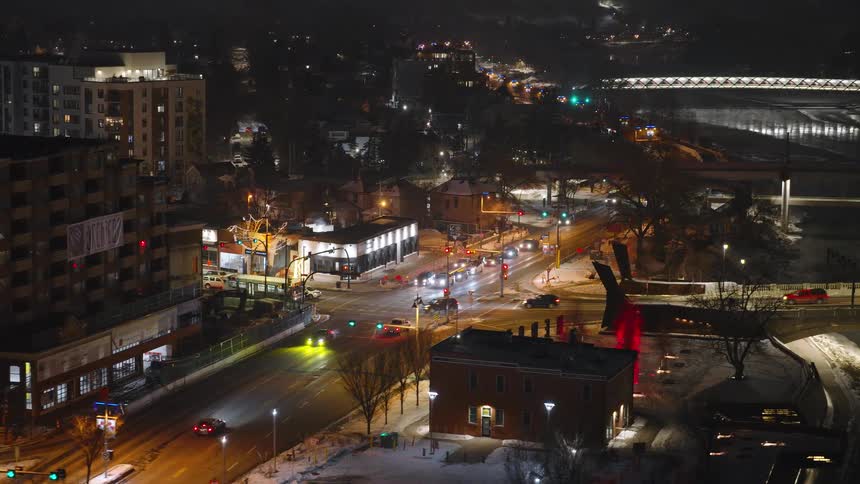 A high angle view shows cars driving through a city intersection at night with street lights and building windows glowing under a dark sky near a river and a bridge