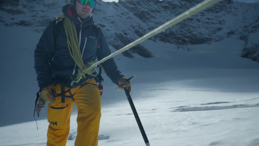 A man wearing a helmet and goggles walks across a snowy mountain slope while holding an ice axe and tethered to a rope during a bright day
