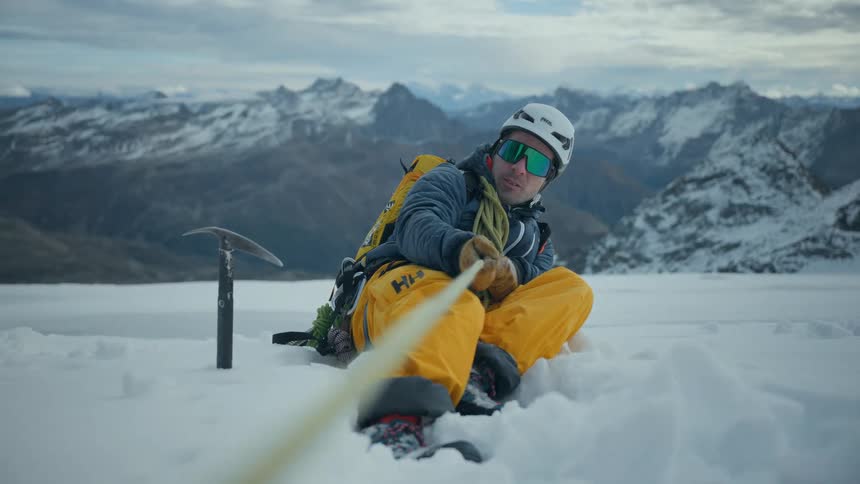 Mountaineer sitting on the snow holding a rope with mountains in the background