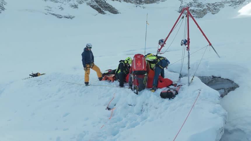 A group of mountain rescuers staged on a snowy glacier next to a deep crevasse they are using a tripod pulley system and specialized gear to carefully tend to an injured person secured in a rescue sled
