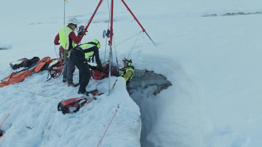A rescue team uses a tripod pulley system to carefully pull an injured person and a rescuer out of a deep glacial crevasse once at the surface the team members stabilize the individual on the snowy ground as they detach the climbing harnesses