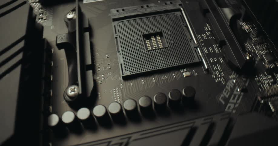 A close view of a dark computer motherboard shows a grid of pins in the central processor socket surrounded by electronic components and circuits