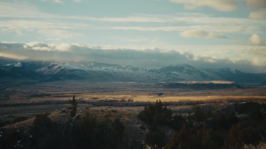 A vast valley with snow capped mountains under a cloudy sky with patches of sunlight