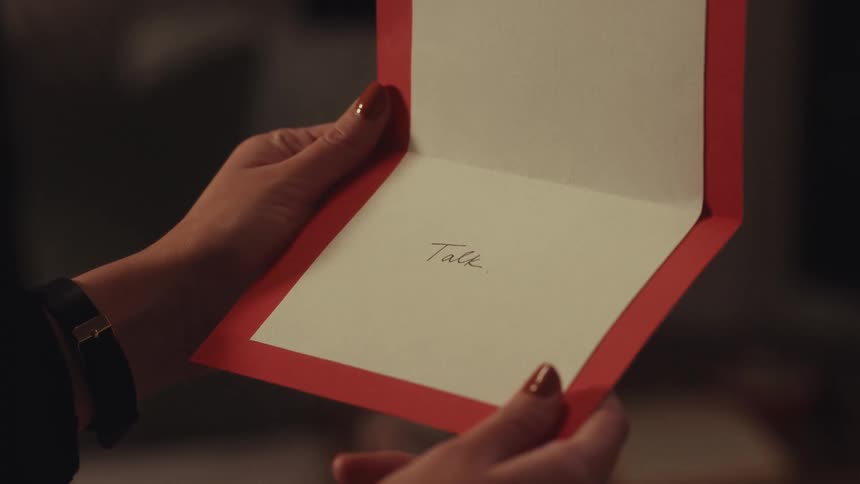 A woman with orange fingernails receives and opens a red greeting card that has the word talk handwritten inside while standing in a dimly lit room