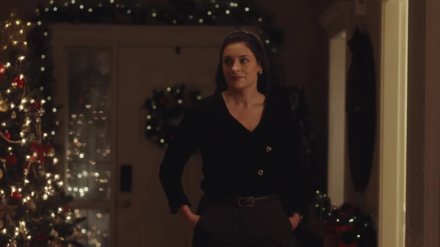 Woman in a black blouse watches another person walk by in a room decorated with lights and a christmas tree