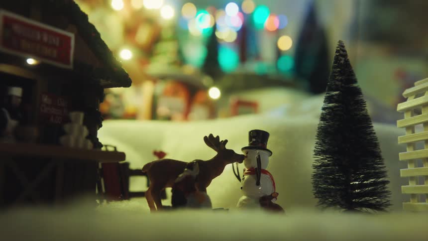 S a miniature christmas village scene featuring a small snowman wearing a top hat standing next to a reindeersoft colorful bokeh lights glow in the background while a tiny evergreen tree and a cider stand frame the festive setting