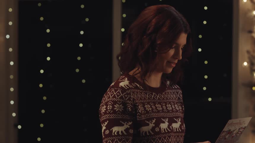 A woman in a festive reindeer sweater opens a red envelope and pulls out a holiday card she reads the card with a thoughtful expression that slowly turns into a warm gentle smile