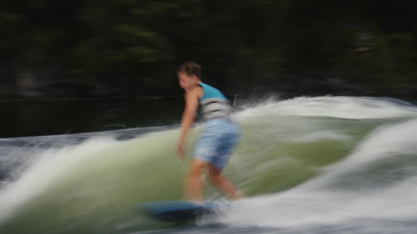 A man in a blue life vest and shorts is wakesurfing on a large churning wave created by a boat he skillfully maintains his balance on the board as he rides the crest of the water near a forested shoreline