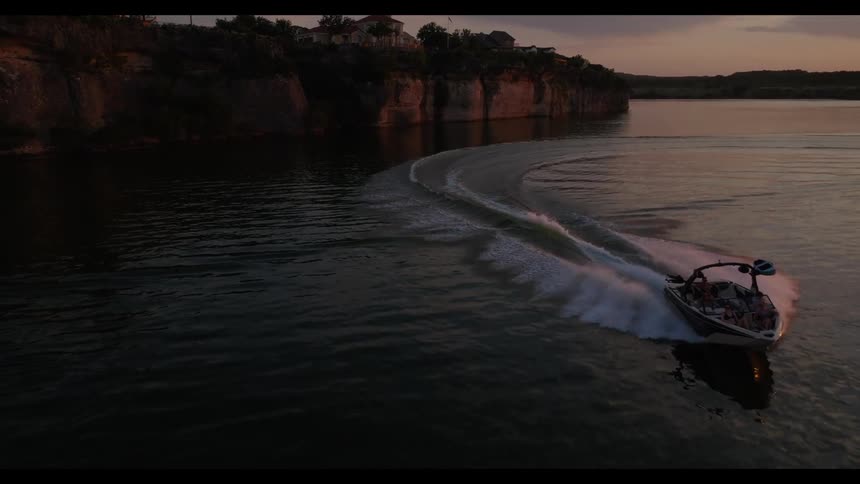 A motorboat carrying a group of friends makes a wide turn on a calm lake at sunset as the wake trails behind it near a rocky cliff with houses under a darkening sky