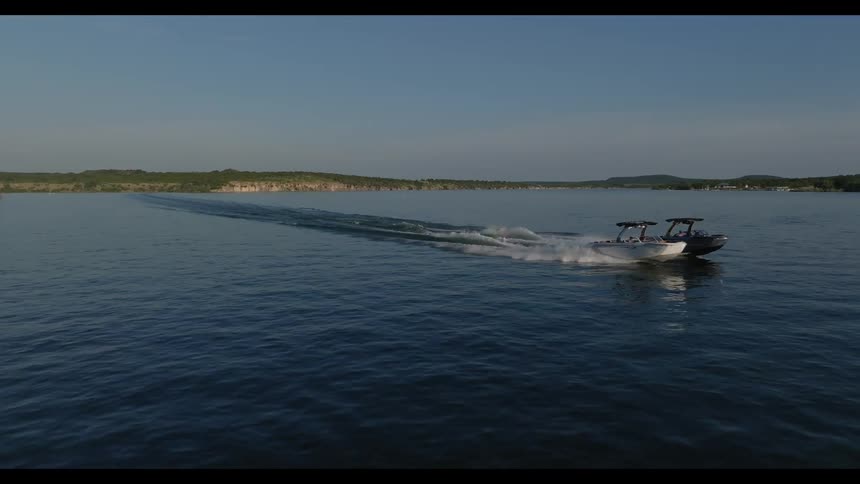 Two motorboats speed across a calm wide body of water leaving long white wakes behind them in the afternoon light the camera captures a wide aerial view of the boats as they pass by a rocky shoreline and green hills in the distance