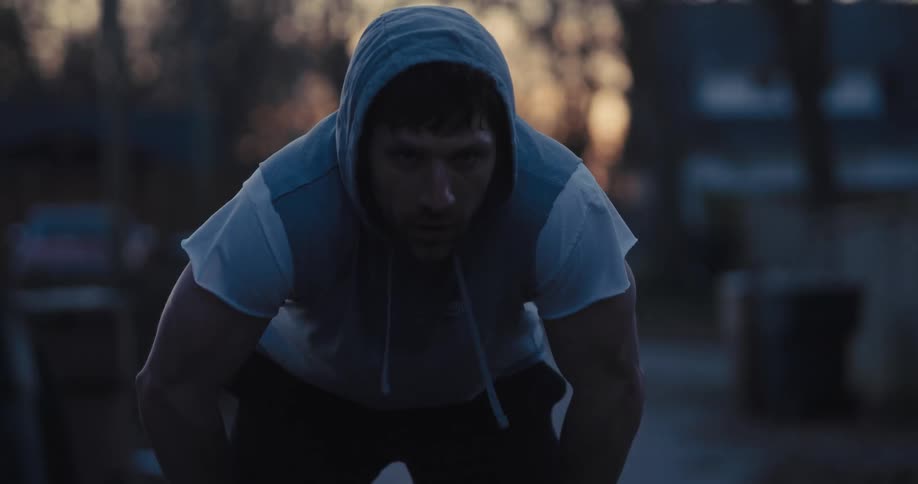 A man wearing a grey hooded vest and a white shirt stands outdoors at dusk while leaning forward with his hands on his knees and looking directly into the camera