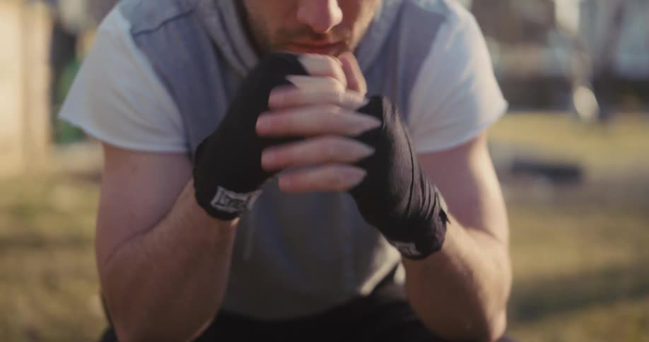 A close view of a man wearing a grey vest and black hand wraps sitting outdoors as he punches his own hand with knuckles and prepares for a workout
