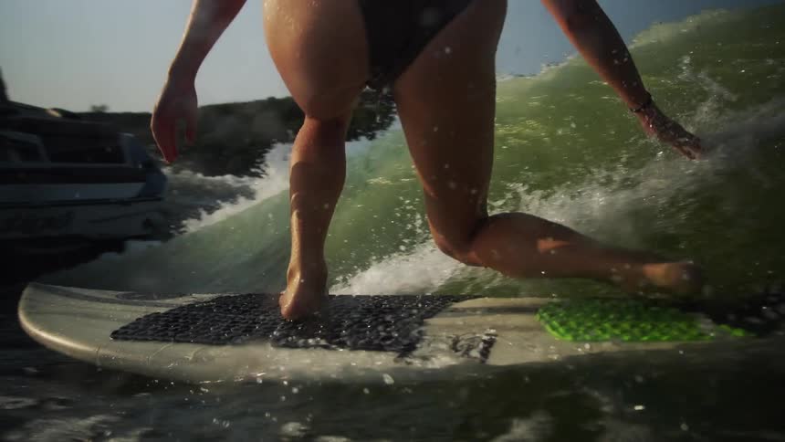 A close view of a woman wearing a dark swimsuit as she balances on a surfboard while riding a large wave behind a motorboat on a lake during a sunny day