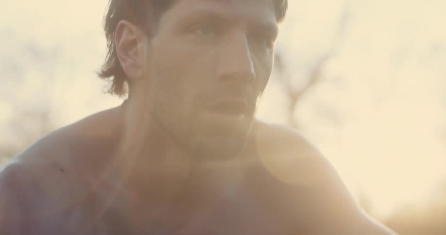 A shirtless young man squatting as he catches his breath after a strenuous exercise session outdoors at sunset