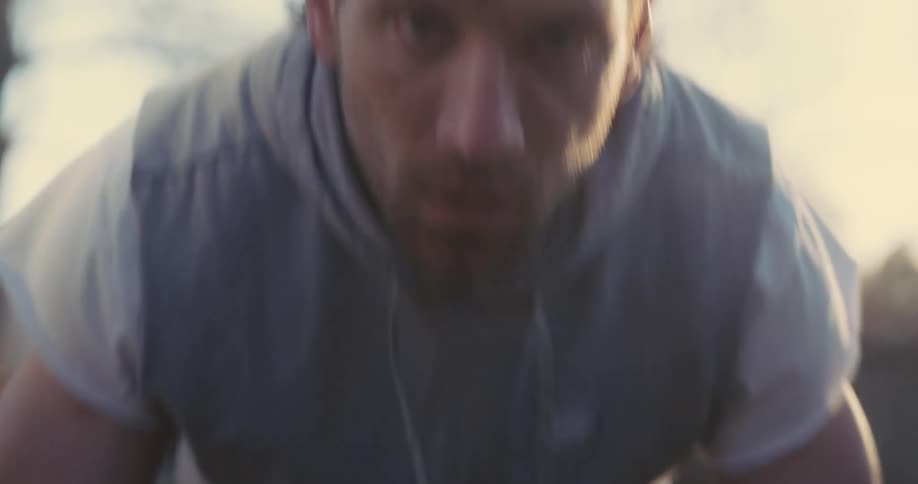 A close view of a man wearing a grey vest and white shirt performing rapid push ups outdoors as he looks directly toward the camera with a focused expression