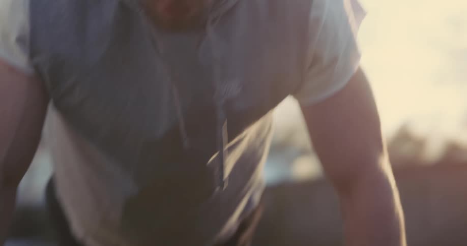 A man wearing a grey vest and black hand wraps performs push ups using a large tire in a grassy yard while the bright sun shines in the background