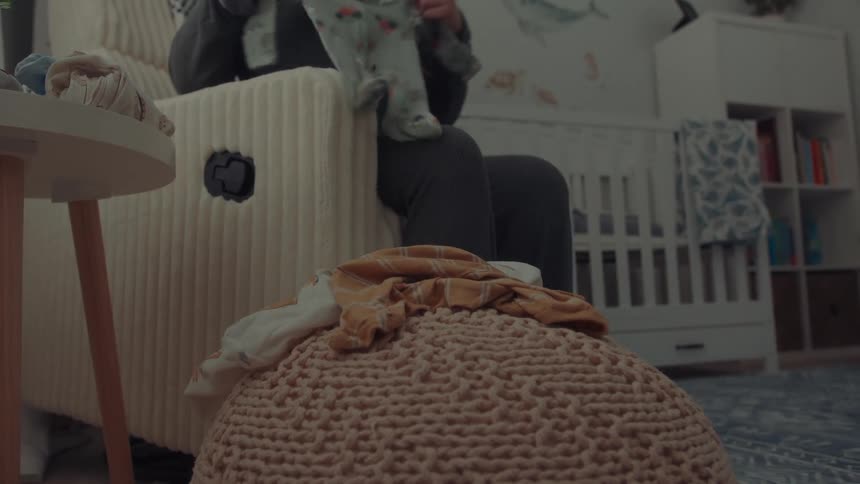 The video shows a person sitting in a nursery chair and sorting through a pile of baby clothes placed on a knitted ottomanthe scene features a soft domestic atmosphere with a white crib and organized shelving visible in the background