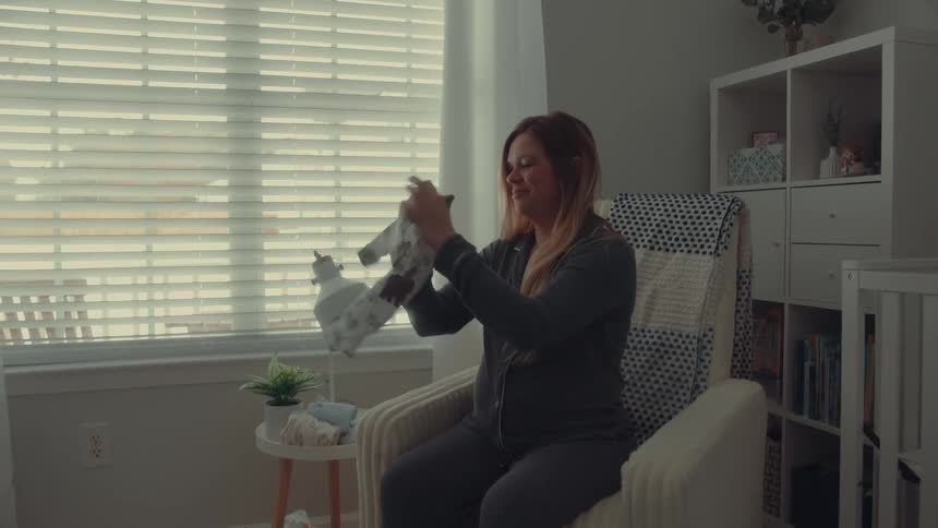 A pregnant woman sitting in an armchair in her bedroom with a baby crib beside her folding baby clothes and placing them on a stool next to the chair