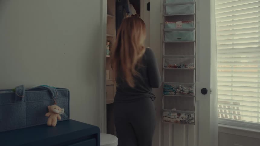 A pregnant woman with long blonde hair organizes baby clothes in a nursery closet she turns away from the closet and walks toward a rocking chair to set down a small garment