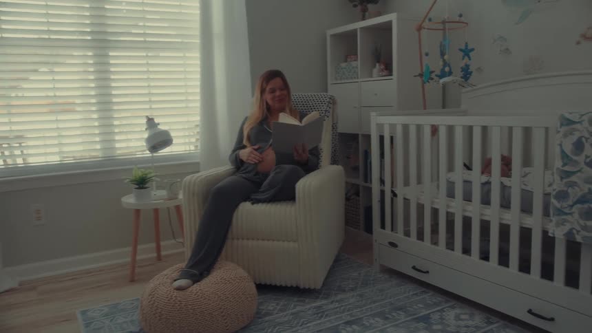 A pregnant woman wearing grey pajamas sits in a cream colored chair inside a nursery and reads a book while resting one hand on her stomach