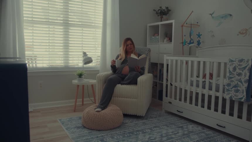 A pregnant woman wearing grey pajamas sits in a cream colored rocking chair in a nursery while reading a book and holding a mug near a white crib