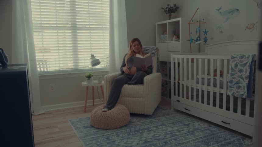 A woman sits comfortably in a rocking chair within a dimly lit nursery quietly reading a bookbeside her a white wooden crib stands prepared on a patterned blue rug surrounded by soft decor and natural light filtering through the blinds