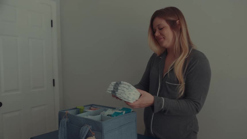 A pregnant woman wearing grey pajamas stands in a nursery and smiles as she organizes stacks of diapers into a blue storage caddy on top of a dresser