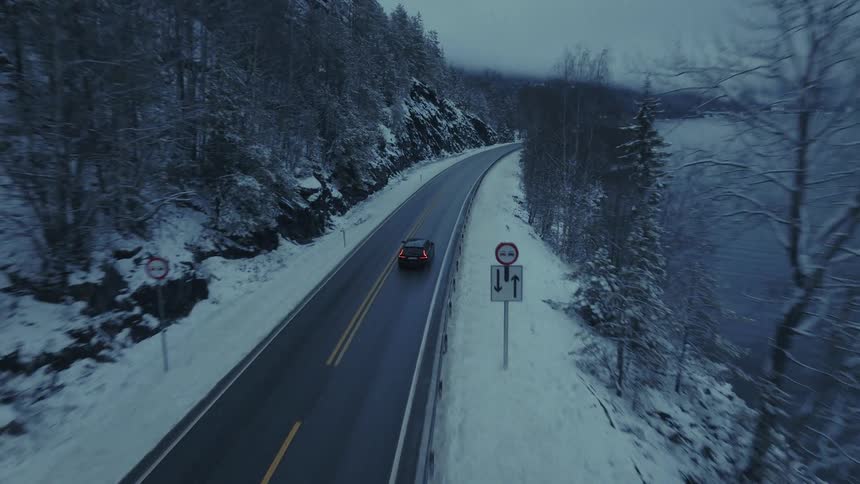 An aerial camera follows a dark car driving along a winding coastal road bordered by steep snowy cliffs and a calm body of waterthe scene is captured in the dim light of dusk or dawn highlighting the contrast between the dark asphalt and the snowcov