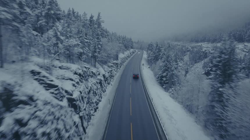 An aerial view shows a dark car traveling down a long wet highway flanked by dense snowcovered pine foreststhe atmosphere is cold and misty with grey skies hanging over the rugged winter landscape
