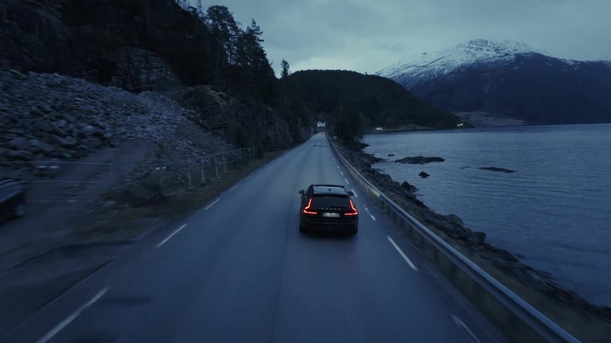 Car driving on a road by a lake with surrounding mountains under a cloudy sky
