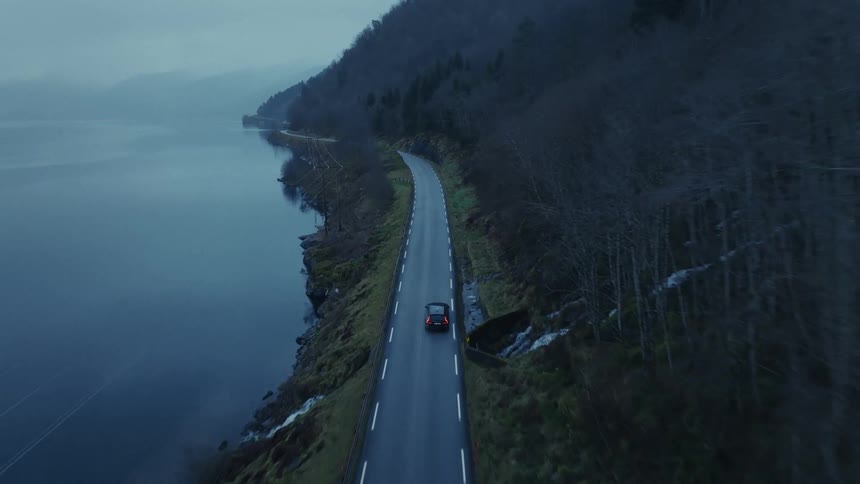 An aerial view shows a car driving on a road that curves between a dark lake and a steep hill with many trees under a grey sky