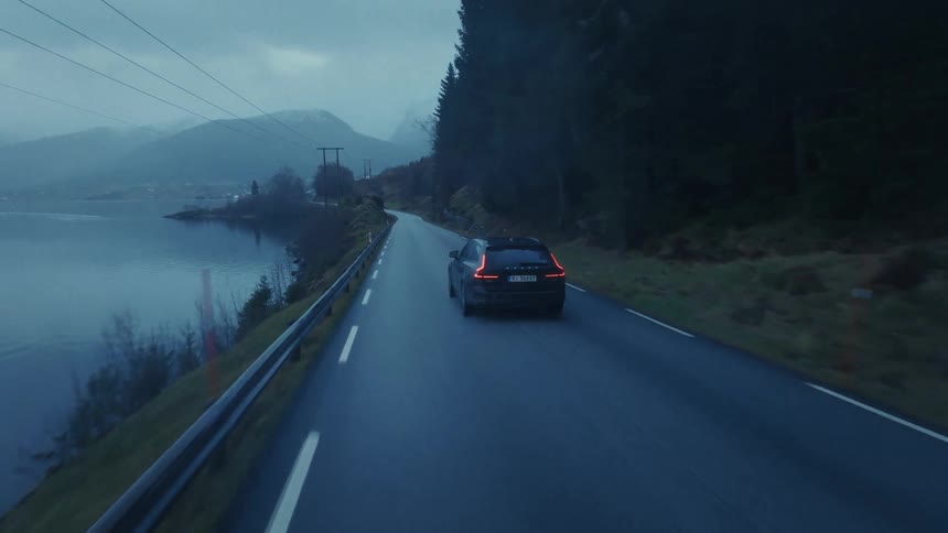 Car drives along winding road beside lake in mountainous landscape at dusk