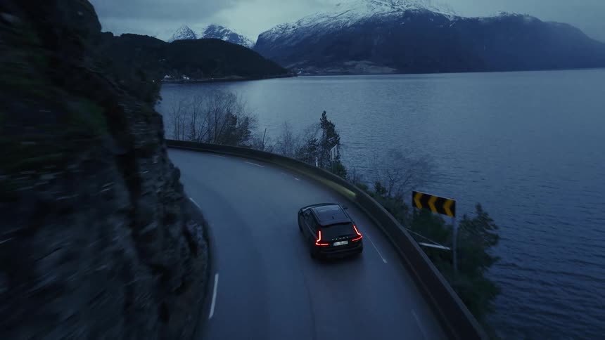 A dark car drives along a winding road on a steep cliffside next to a large body of water with snow covered mountains visible in the distance under a dim evening sky